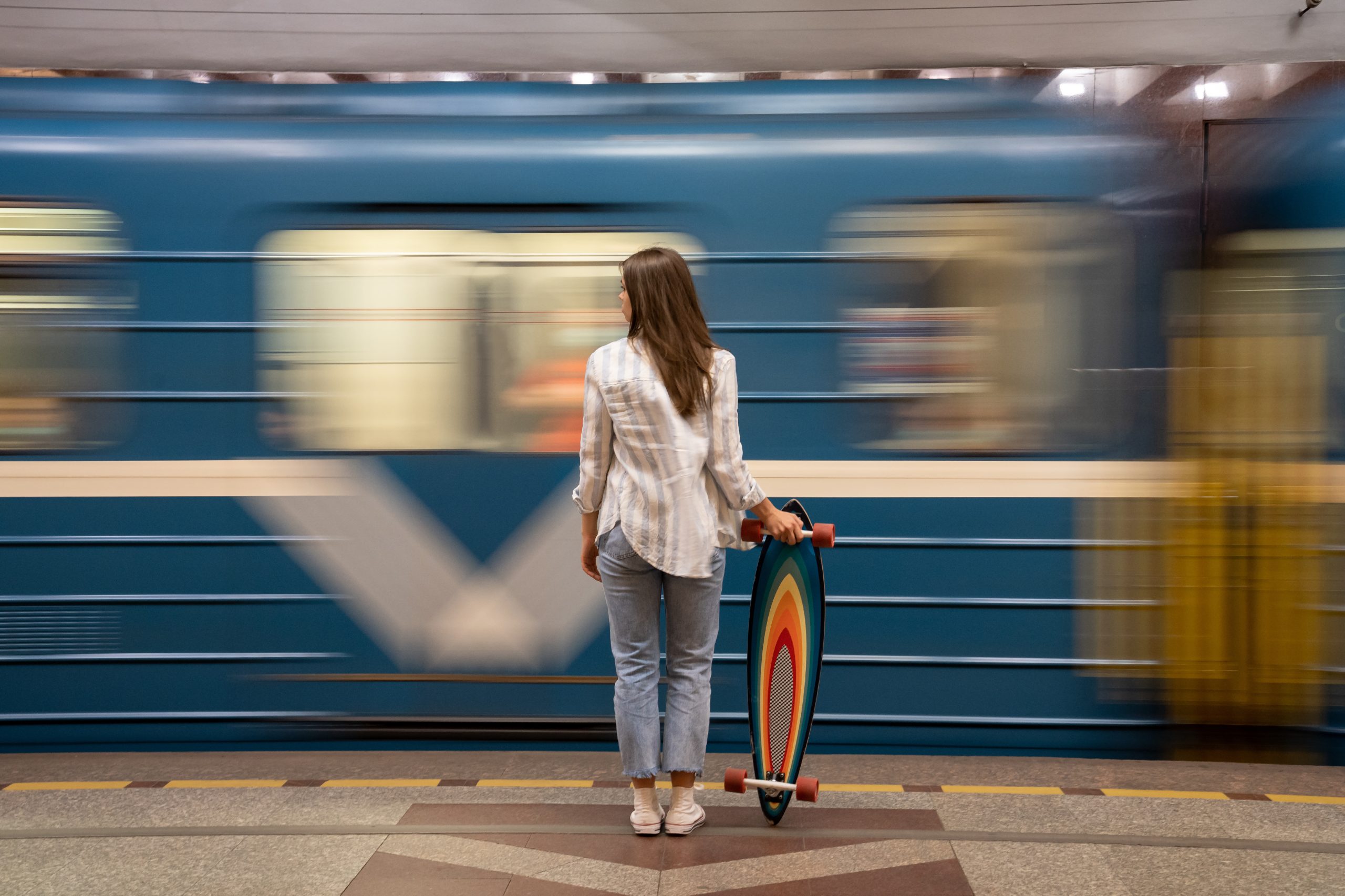 Girl with longboard standing on underground platform with blurry moving blue train on background